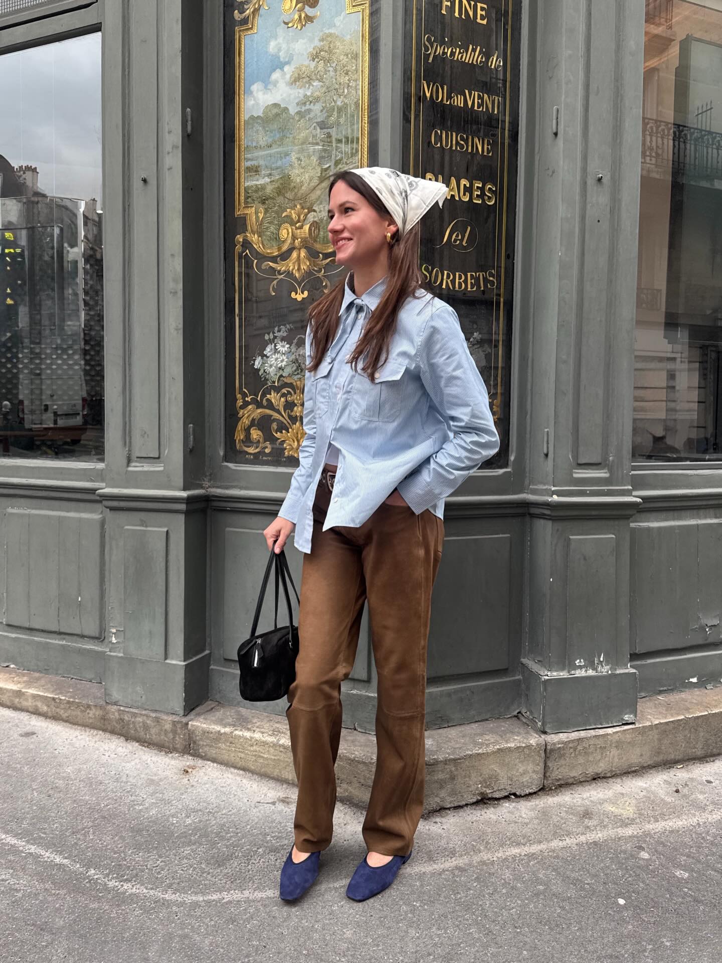 An image of an influencer wearing a white bandana, light blue button-down shirt, brown suede trousers and navy suede ballet flats in Paris