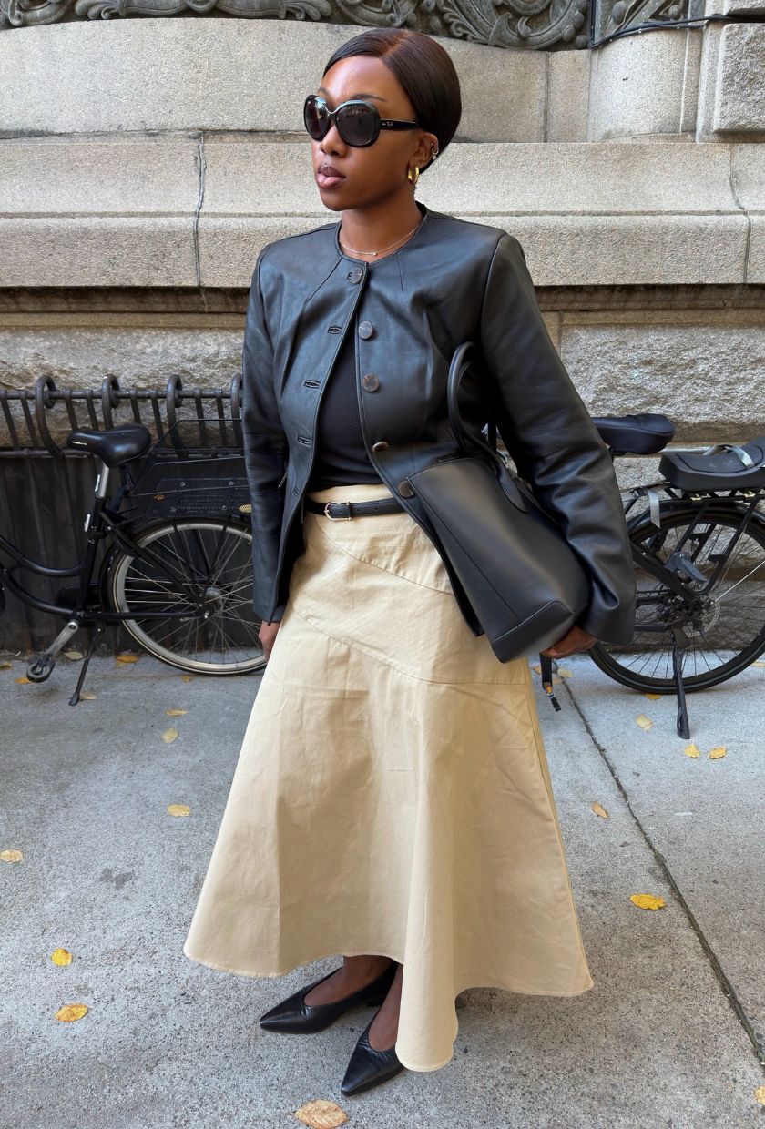 @nnennaechem wearing a black leather waisted blazer, black t-shirt, cream a-line midi skirt, black tote bag, pumps and sunglasses