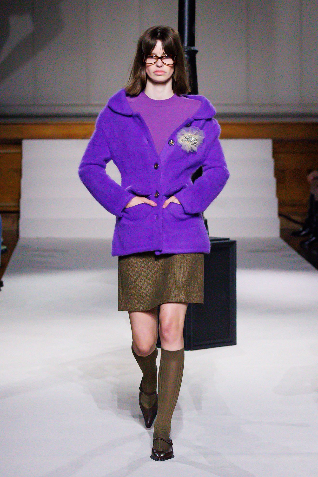 a model on the Kent and Curwen runway wears a purple t-shirt, cardigan and brown skirt and knee high socks with pointed flats.