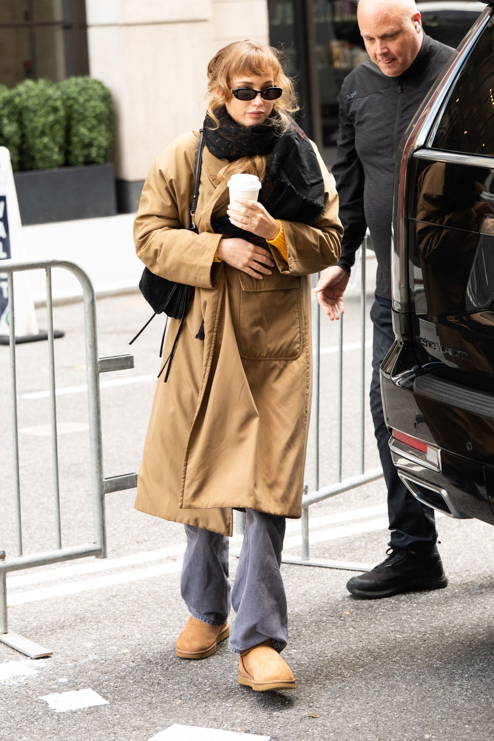 Jennifer Lawrence steps outside in New York wearing a beige trench coat with a black scarf, black sunglasses, grey jeans and Uggs.