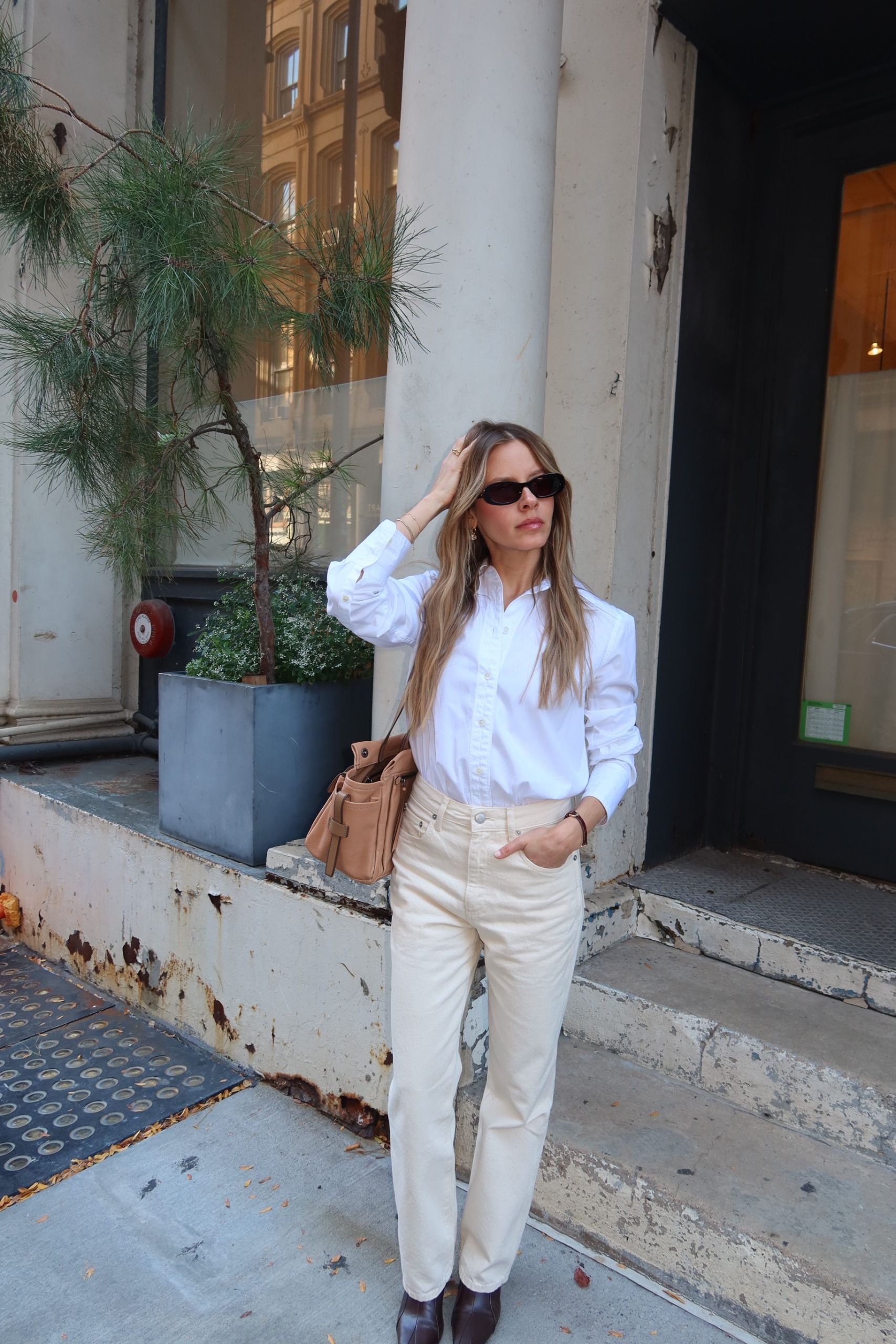 nikki wears a white button-down shirt, ivory jeans, brown boots and a tan bag.