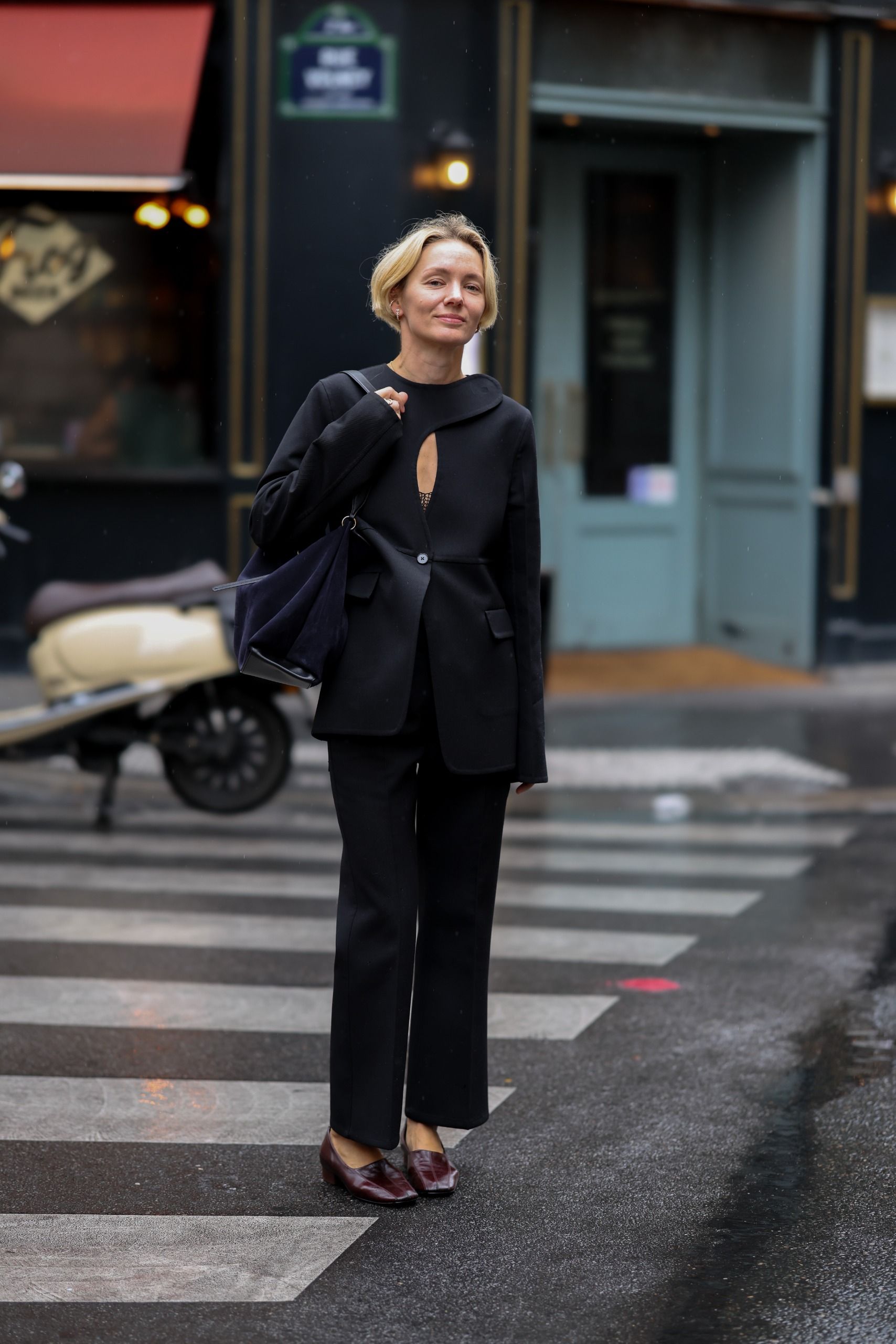 Yana McKillop wearing a black velvet jacket, black straight leg trousers; burgundy leather low heel slingback shoes; and a dark navy shoulder bag; carrying a navy bag