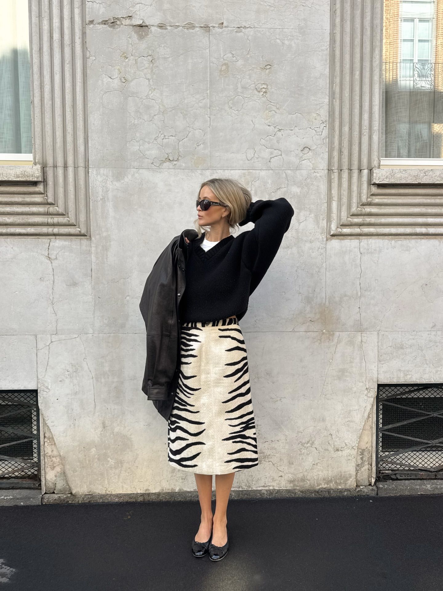An image of an influencer wearing a zebra print midi skirt, a black jumper, black ballet flats and a black leather jacket