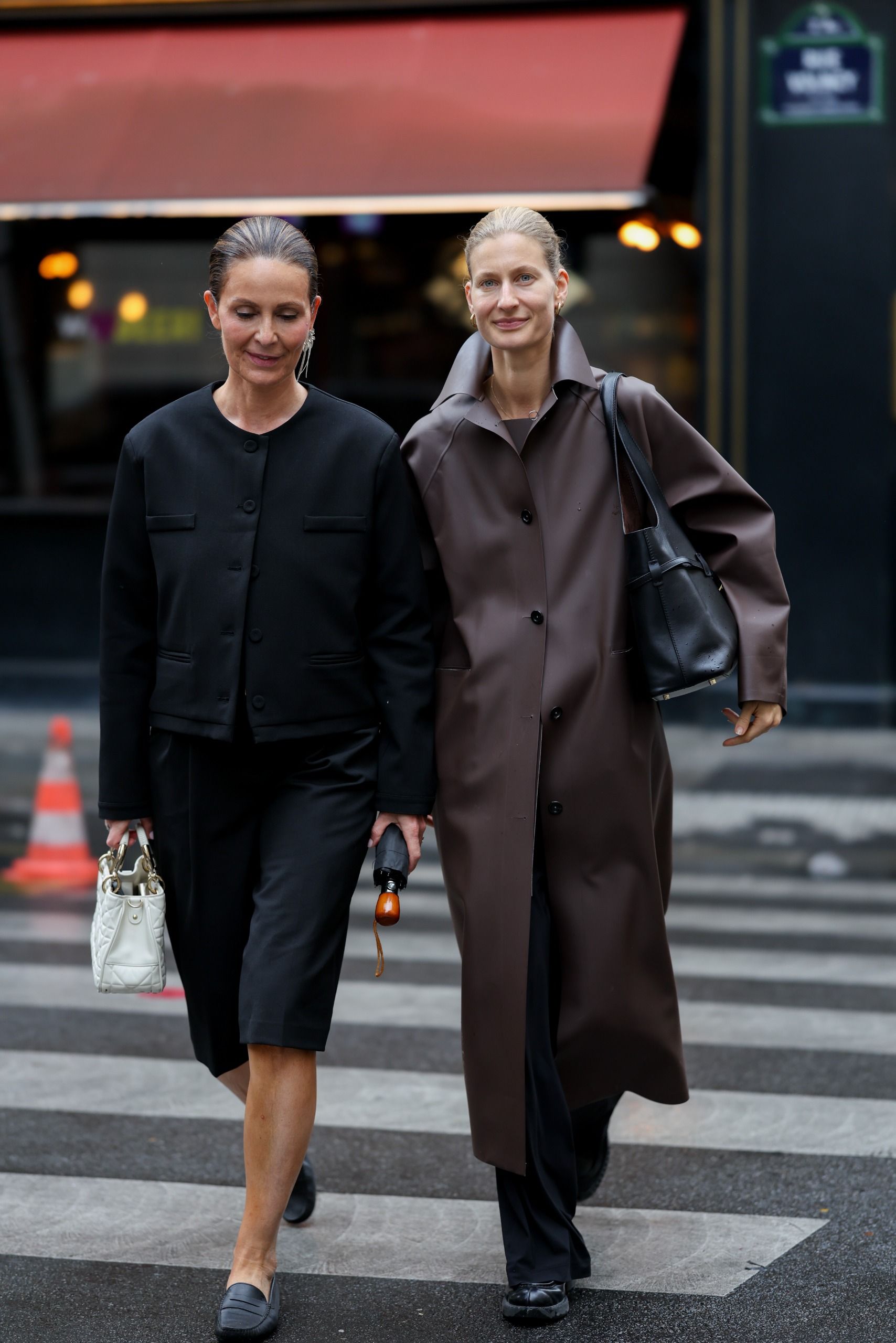 Miluska Van't Lamnwearing a black collarless jacket; black tailored shorts; leather loafers; a white Dior bag; Simone Sniekers is wearing a brown long coat; black wide leg trousers; chunky loafers