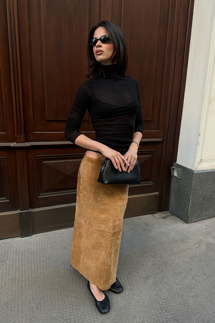 A woman wearing a black turtleneck sweater with a tan suede maxi pencil skirt, black ballet flats, and a black clutch bag.