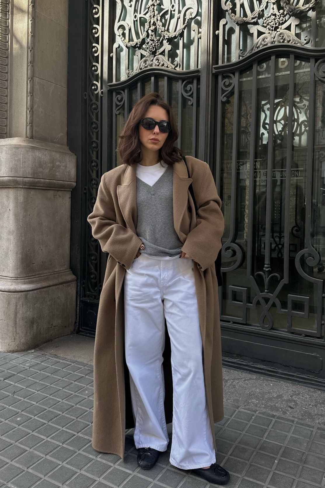 Woman wears camel coat, grey v-neck jumper, white T-shirt, white jeans and black loafers