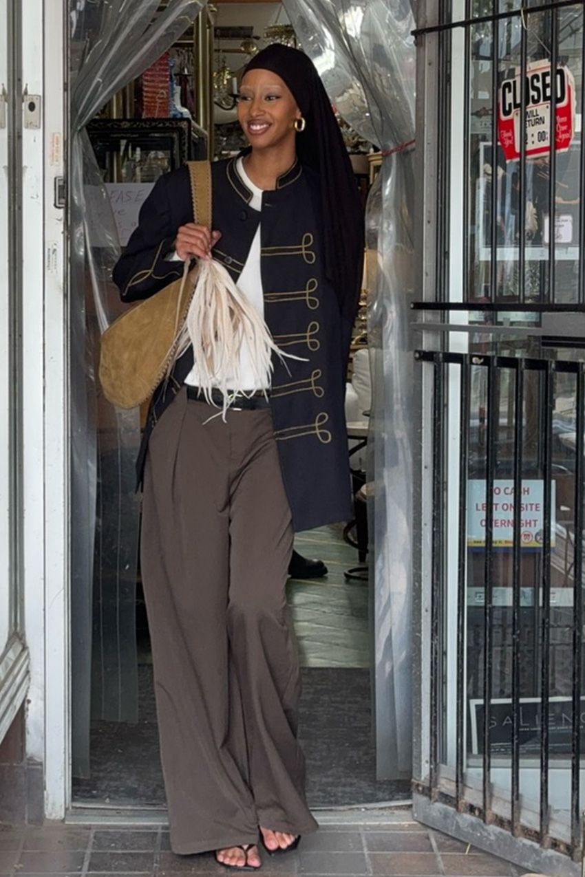 A woman wearing a navy Napoleon jacket with a white blouse, brown wide-leg trousers, black sandals, and a suede slouchy bag.
