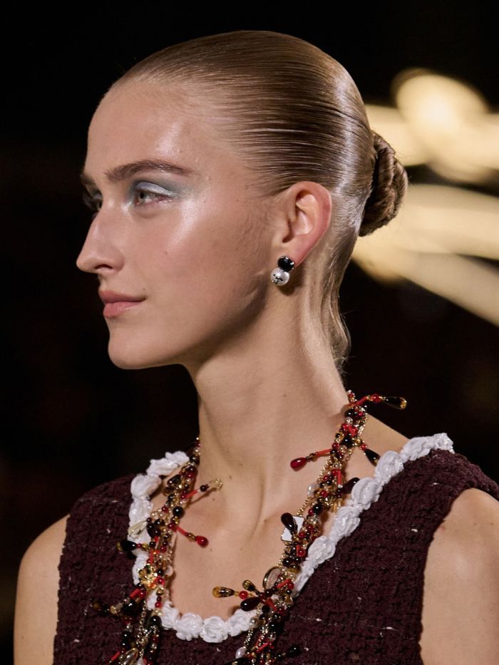 A model walking the runway at the S/S 27 Chanel show with a slicked-back bun and glowing skin
