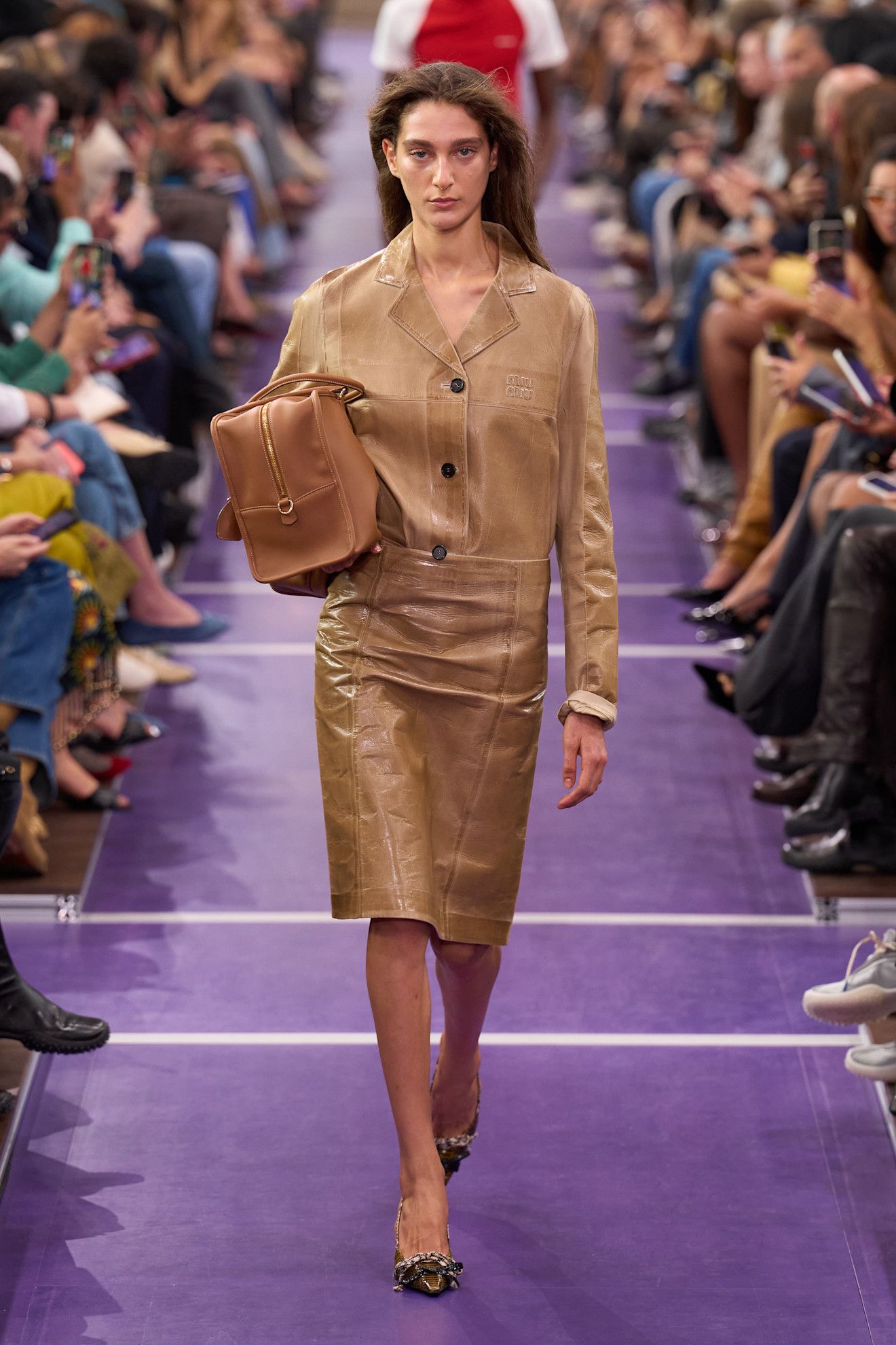 A Miu Miu spring/summer 2027 runway model wearing a leather jacket tucked into a leather pencil skirt styled with pumps and a bowler bag.