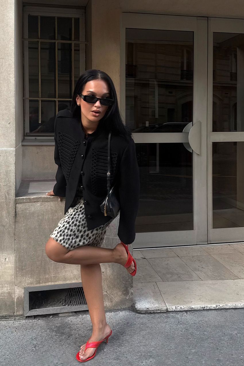 A woman standing outside wearing a black band jacket with a leopard print pencil skirt, red thong sandals, and a black shoulder bag.