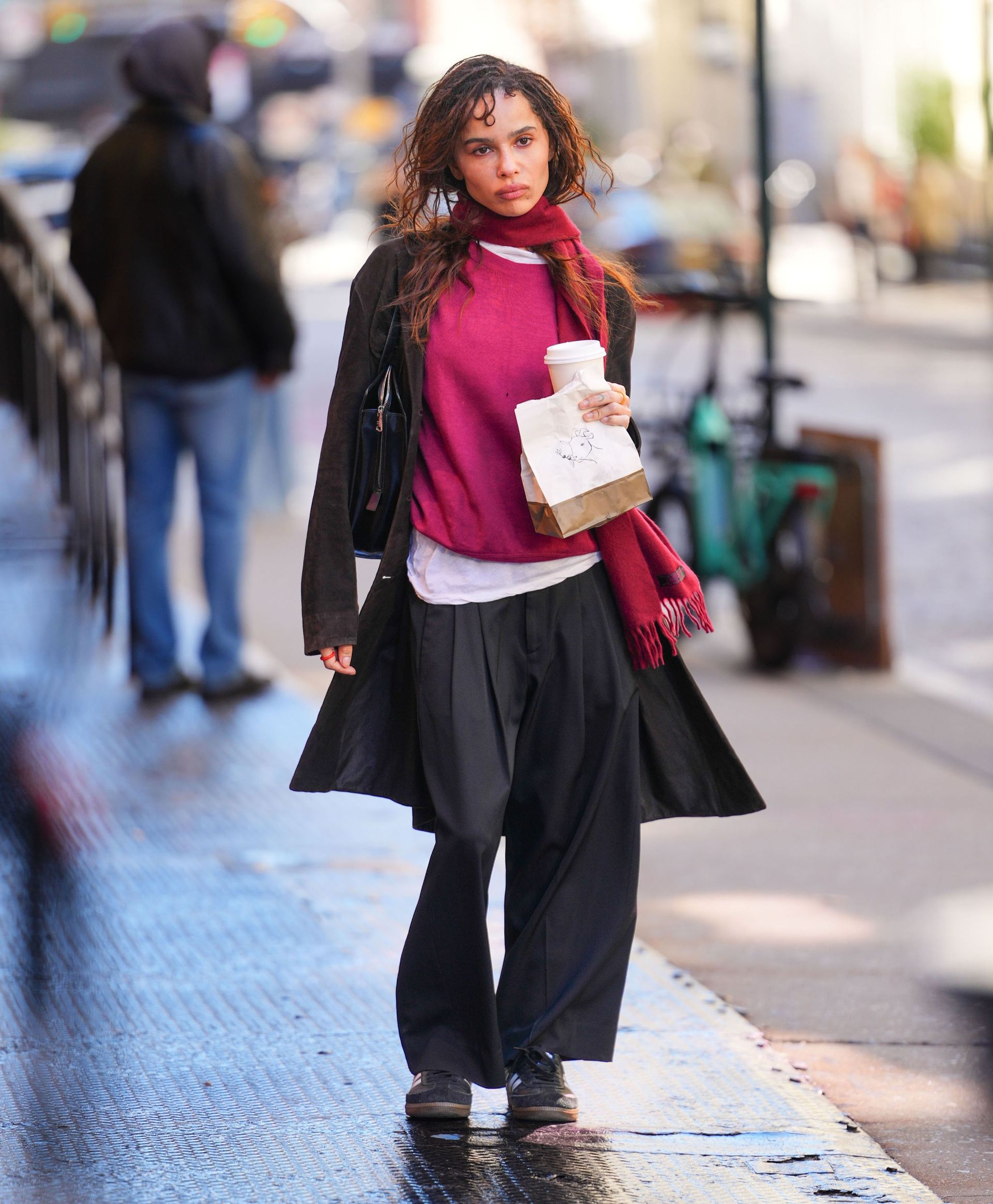 Zoe Kravitz walks down the street in New York wearing black trousers with black trainers, a black jacket and a raspberry jumper and scarf.