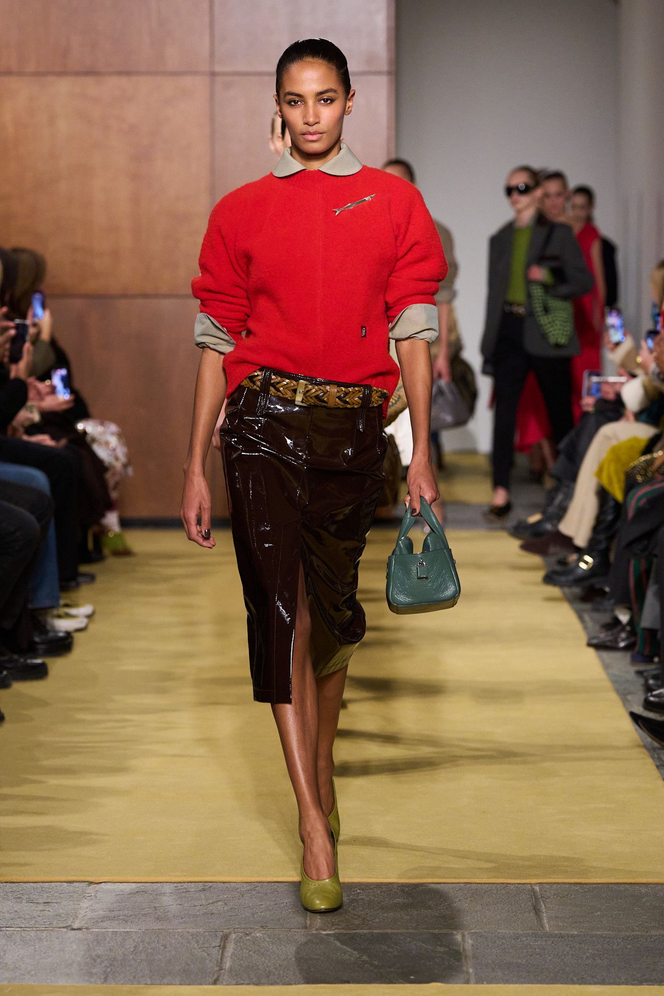 A Tory Burch fall/winter 2026 runway model wearing a tan button-down and red crewneck sweater with a leather pencil skirt and pumps.