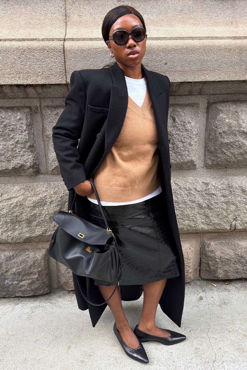 A woman wearing a long black coat over a tan V-neck sweater and white T-shirt, styled with a leather pencil skirt, ballet flats, and a bag.