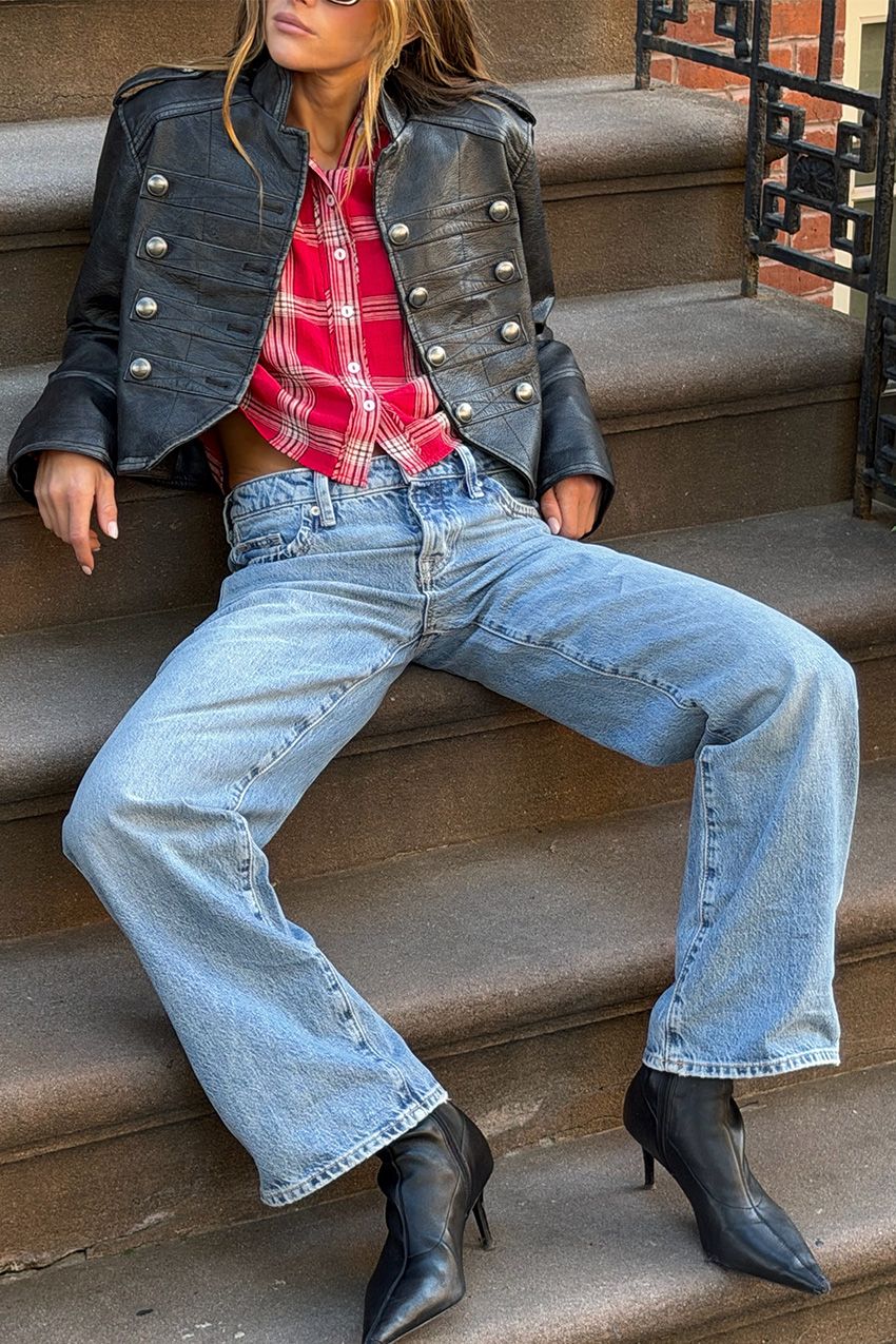 A woman sitting on steps outside wearing a leather military jacket with a red flannel shirt, light wash jeans, and black ankle boots.