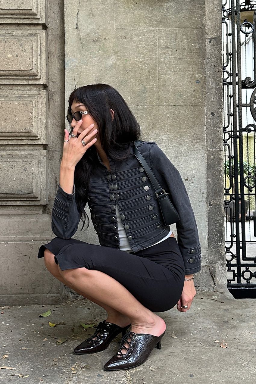 A woman wearing a black military jacket with a white t-shirt, black capris, black heeled mules, and a black shoulder bag.