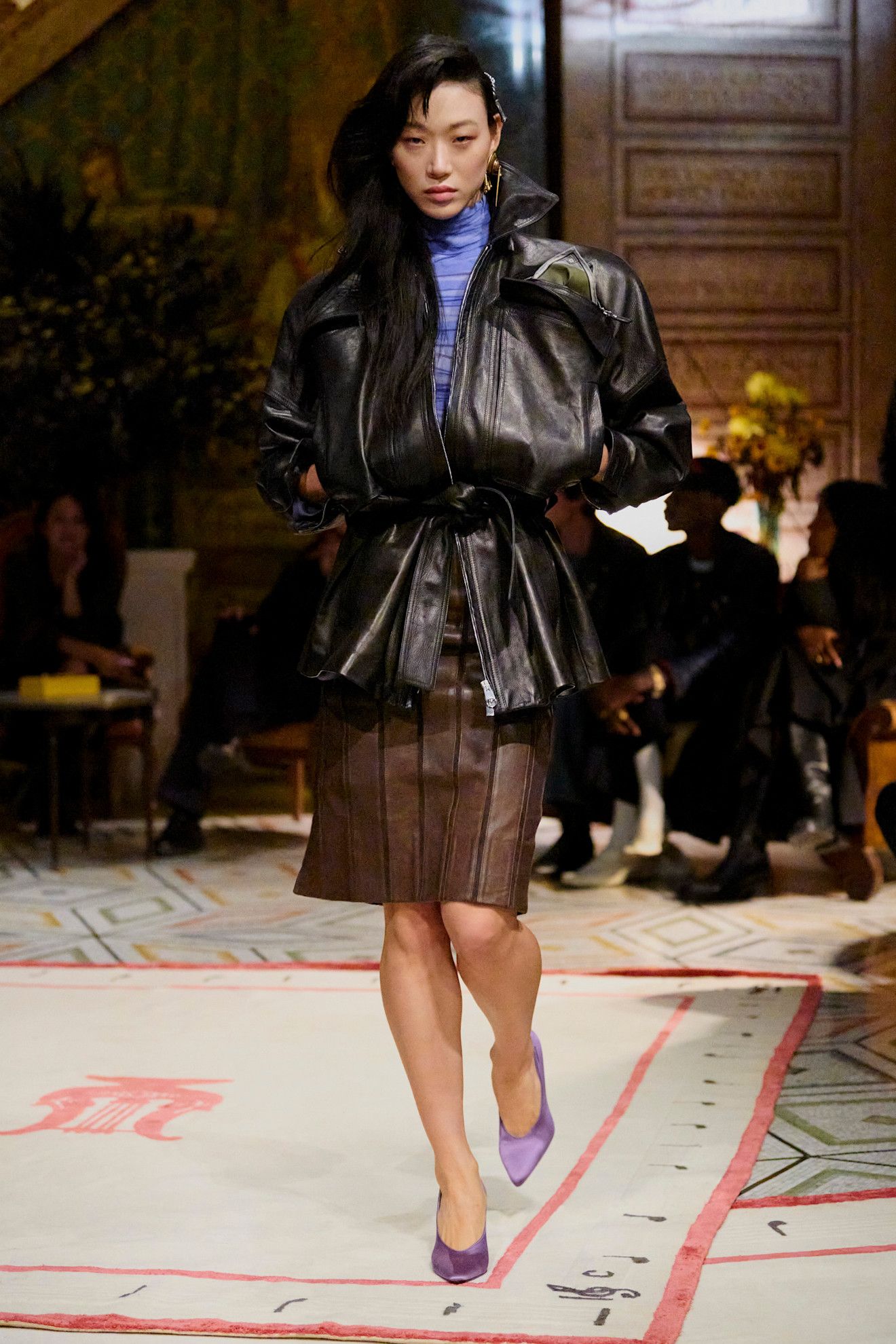 A Versace spring/summer 2026 runway model wearing a black leather bomber styled with a purple shirt, brown paneled leather skirt, and purple pumps.