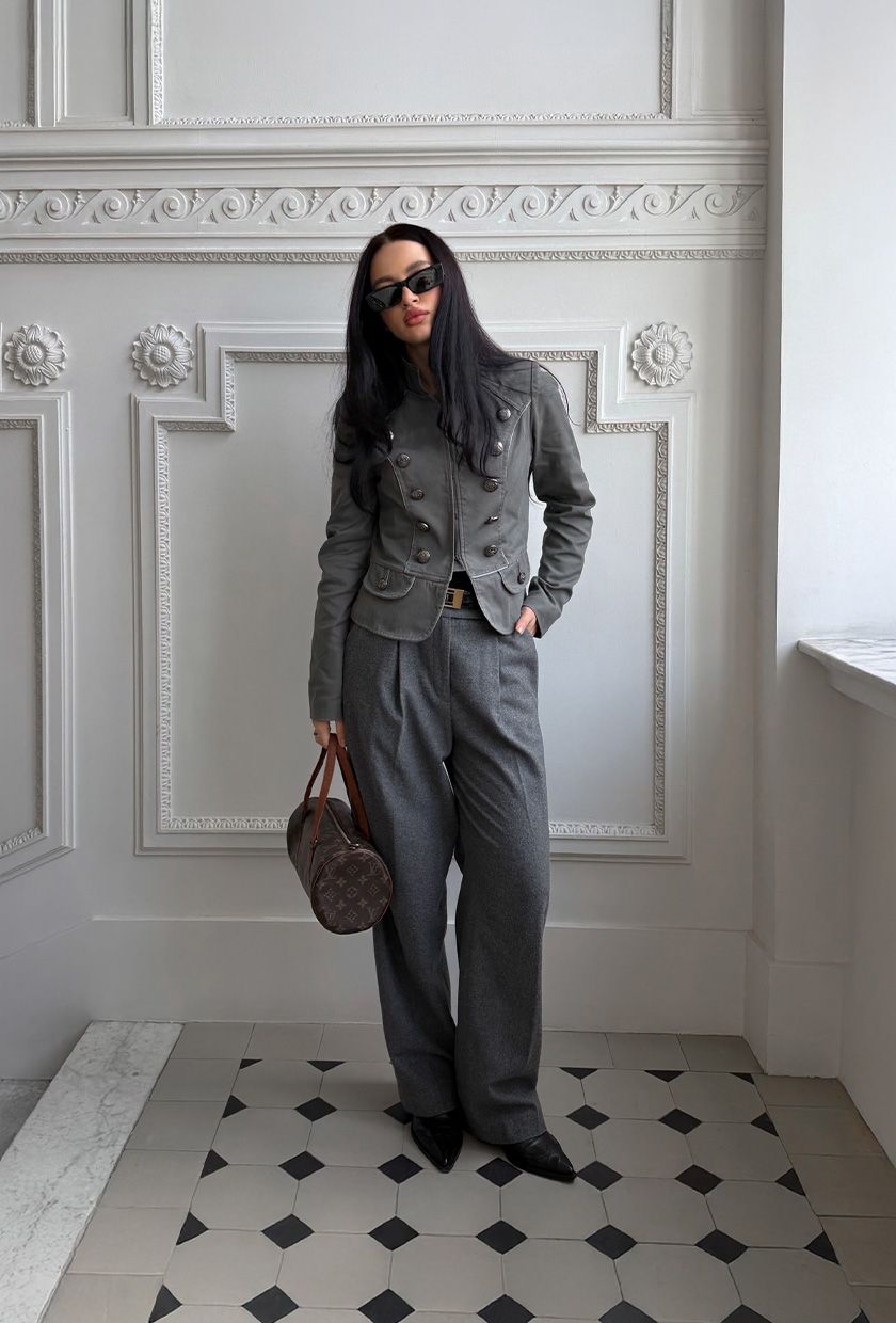A woman standing in the hallway wearing a gray military jacket with gray wool trousers, pointed heels, and a Louis Vuitton bag.