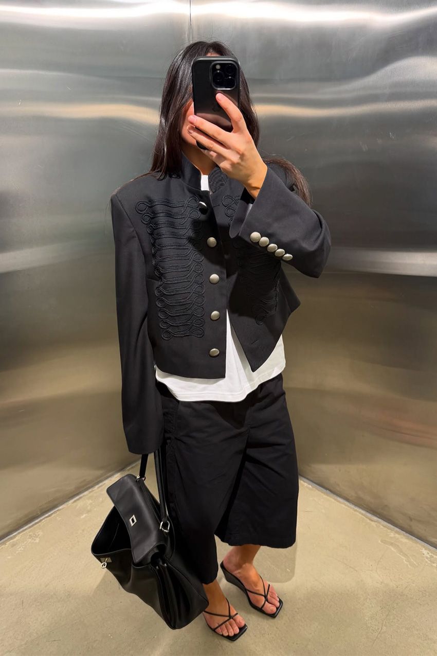 A woman standing in an elevator wearing a black jacket with a white t-shirt, black baggy jean shorts, and black wedges.