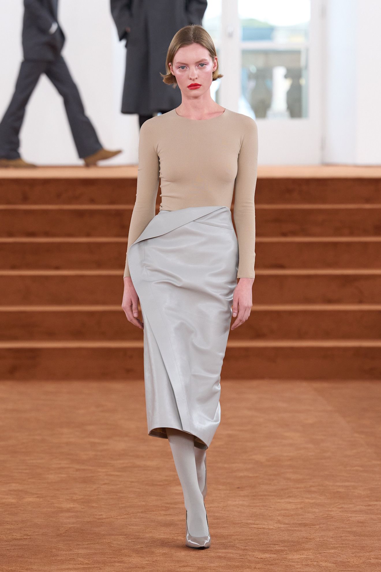 A Jil Sander spring/summer 2026 runway model wearing a tan T-shirt with a gray pencil skirt, gray tights, and gray pumps.