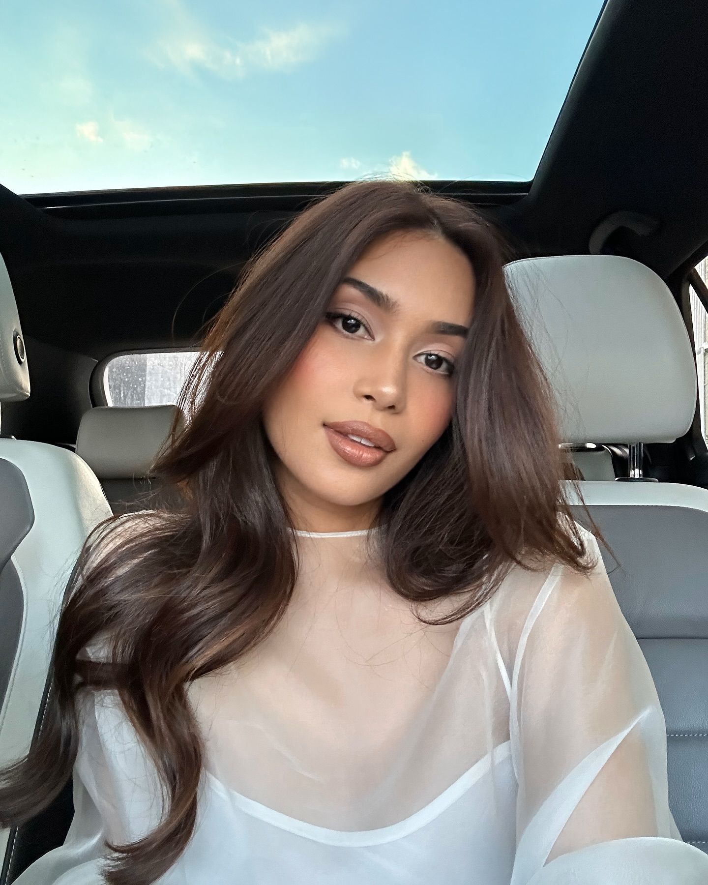 A woman taking a selfie from a car wearing a white dress.