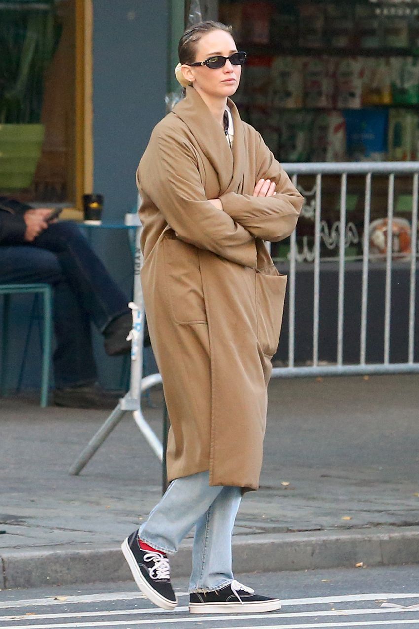 Jennifer Lawrence in NYC wearing a camel coat, large pendant necklace, white T-shirt, jeans, and Vans sneakers