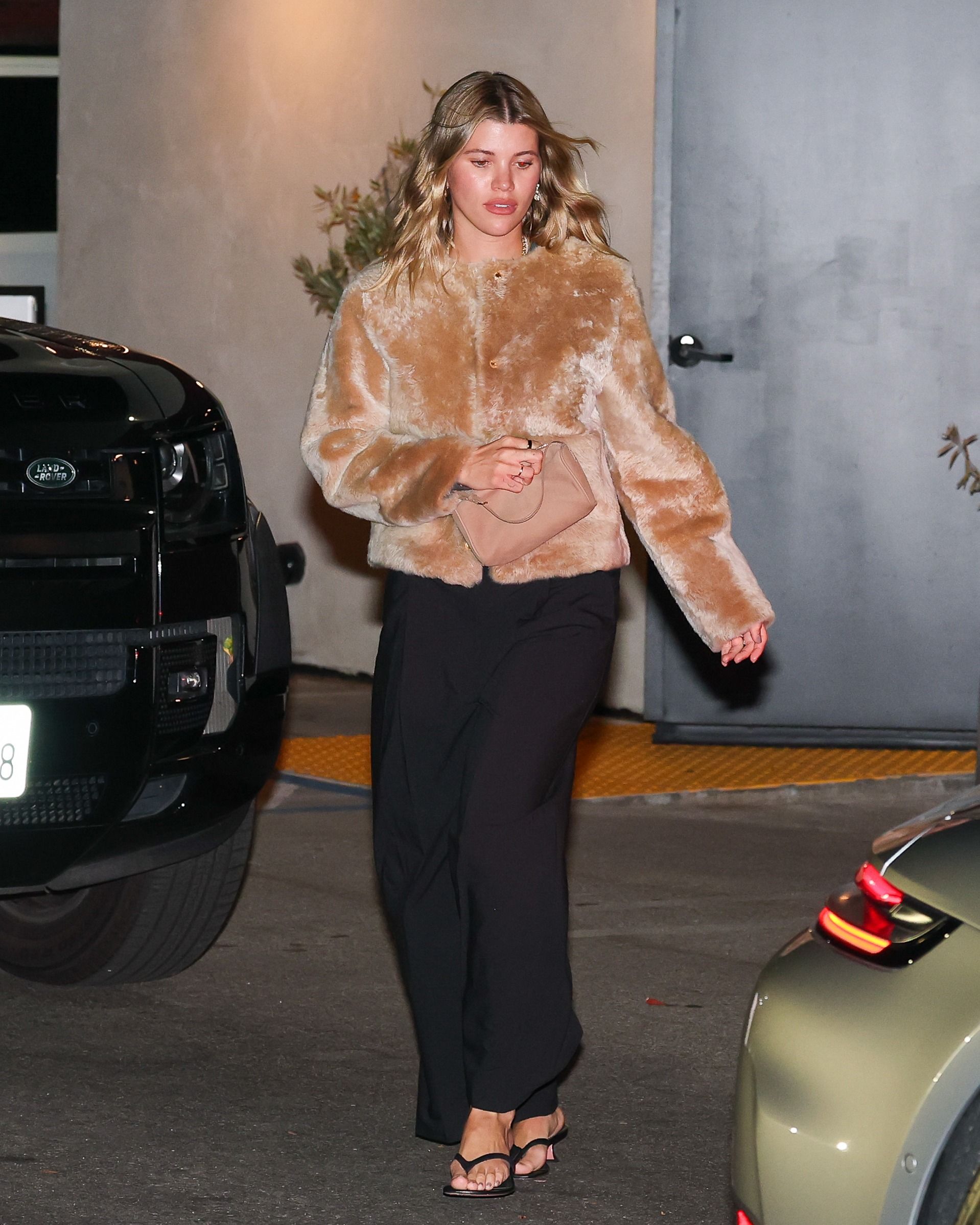 Sofia Richie steps out wearing a faux fur jacket with black trousers and black kitten heel flip-flops. She carries a beige bag in her hands.
