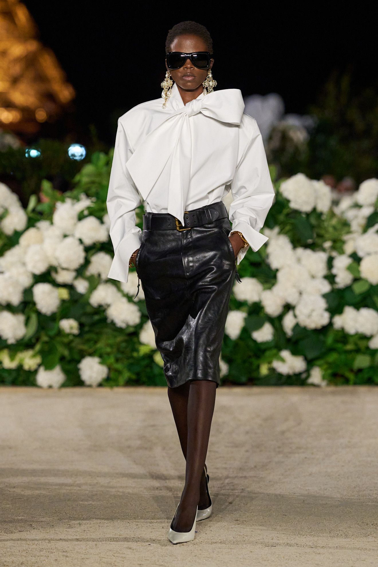 A Saint Laurent spring/summer 2026 runway model wearing a white bow blouse with a black leather pencil skirt, sheer tights, and pointed heels.