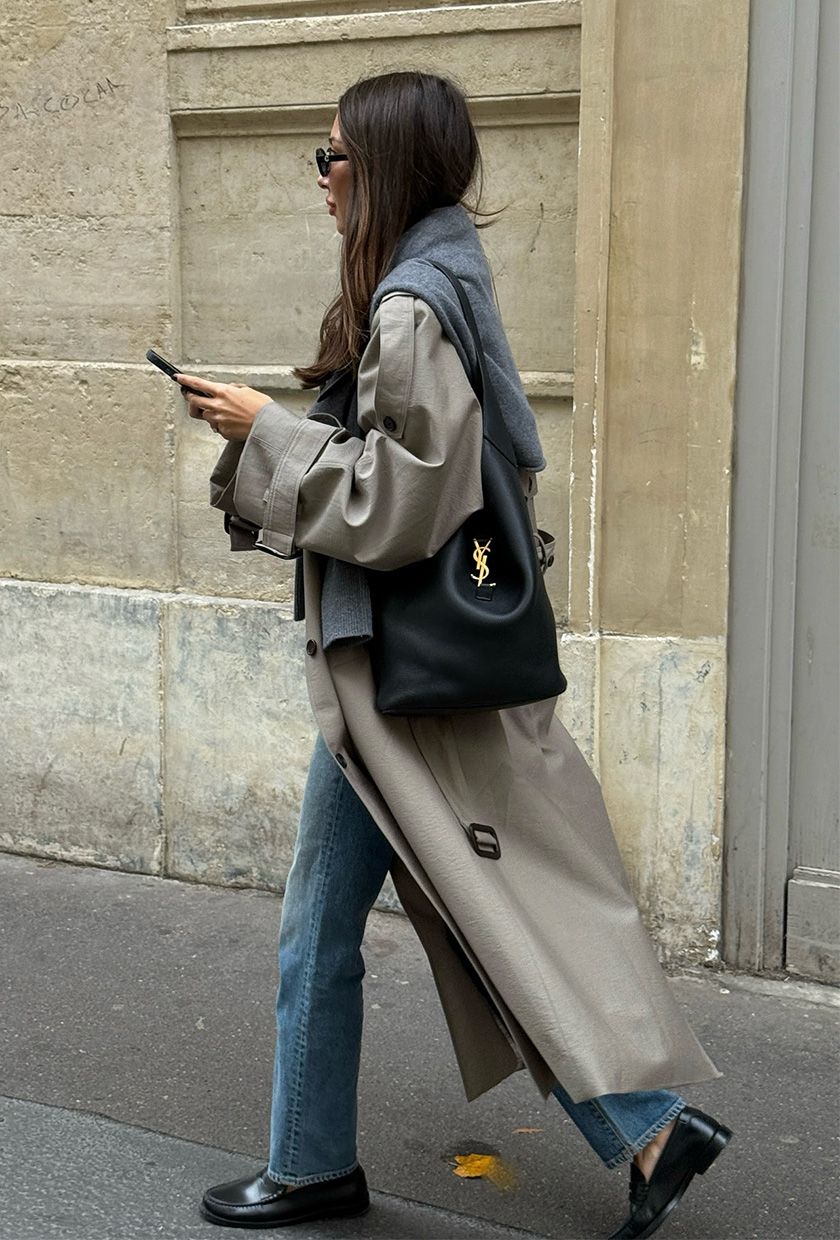 A woman walking down the street wearing a trench coat with a gray sweater around the shoulders, straight jeans, black loafers, and a black slouchy bag.