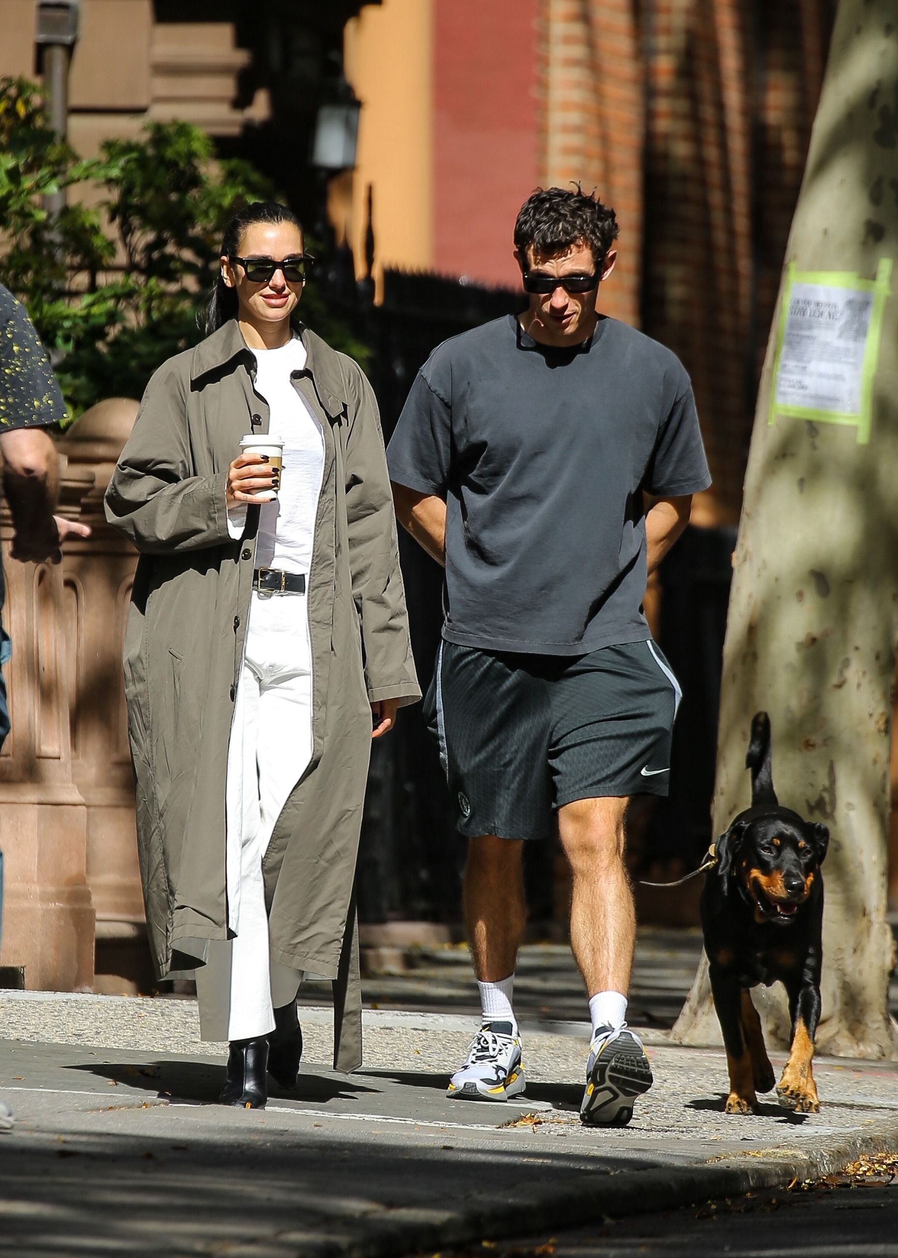 Dua lipa wears a trench coat, white tee, black belt, white jeans, and black heeled boots.