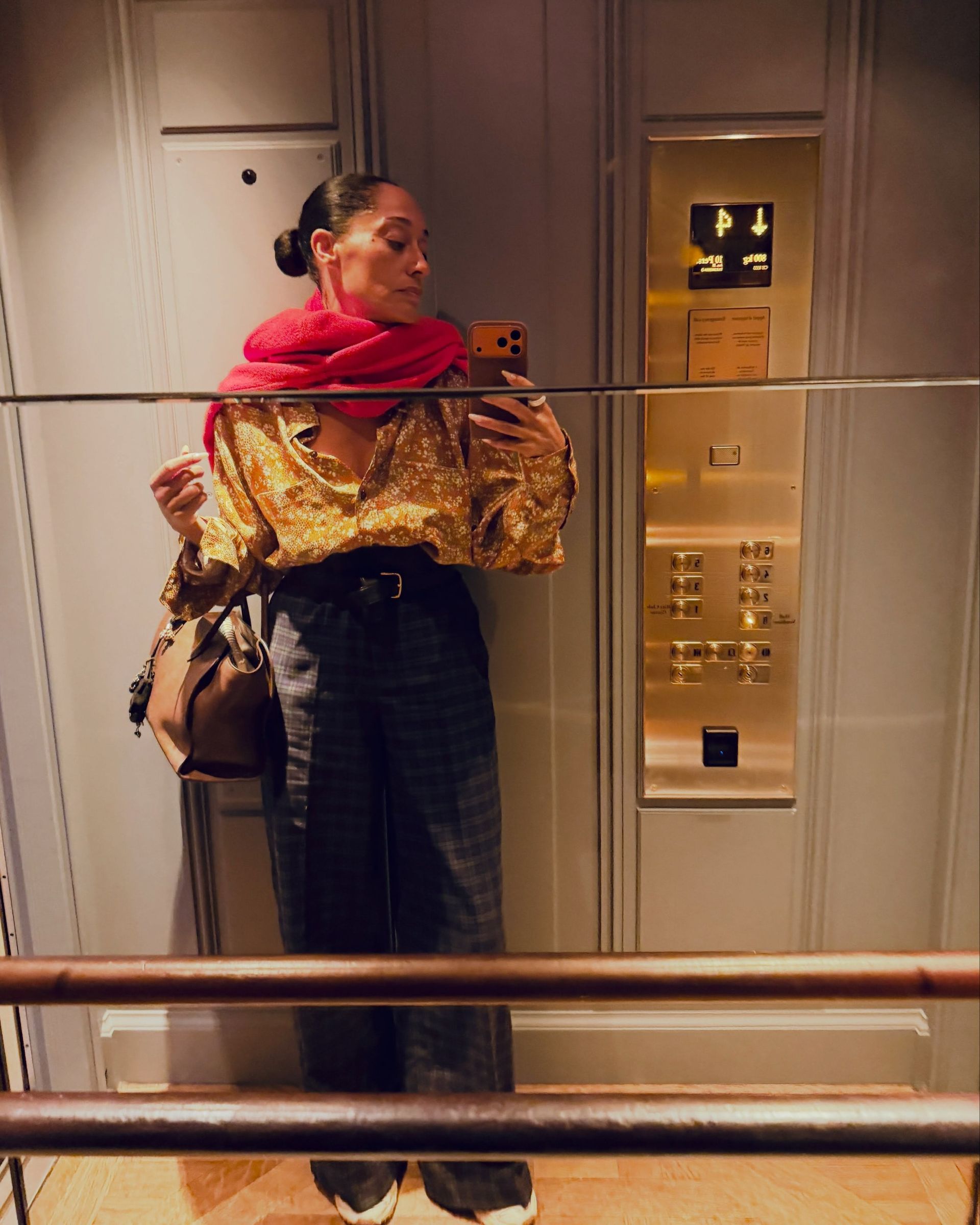 Tracee Ellis-Ross takes a mirror selfie wearing a gold button-down shirt with a pink scarf and checked trousers.
