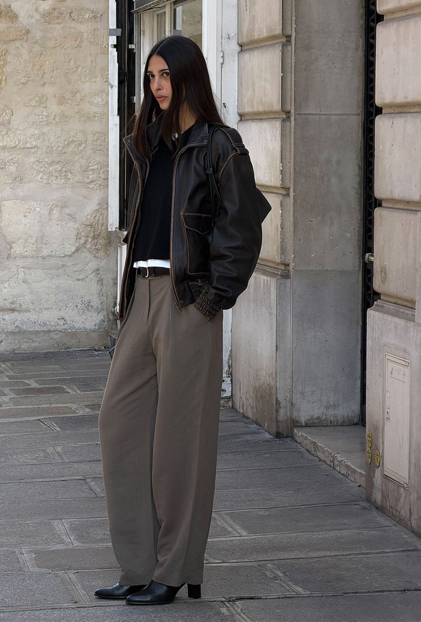 A woman wearing a brown leather bomber jacket over a black sweater and white T-shirt, styled with gray trousers and black ankle boots.