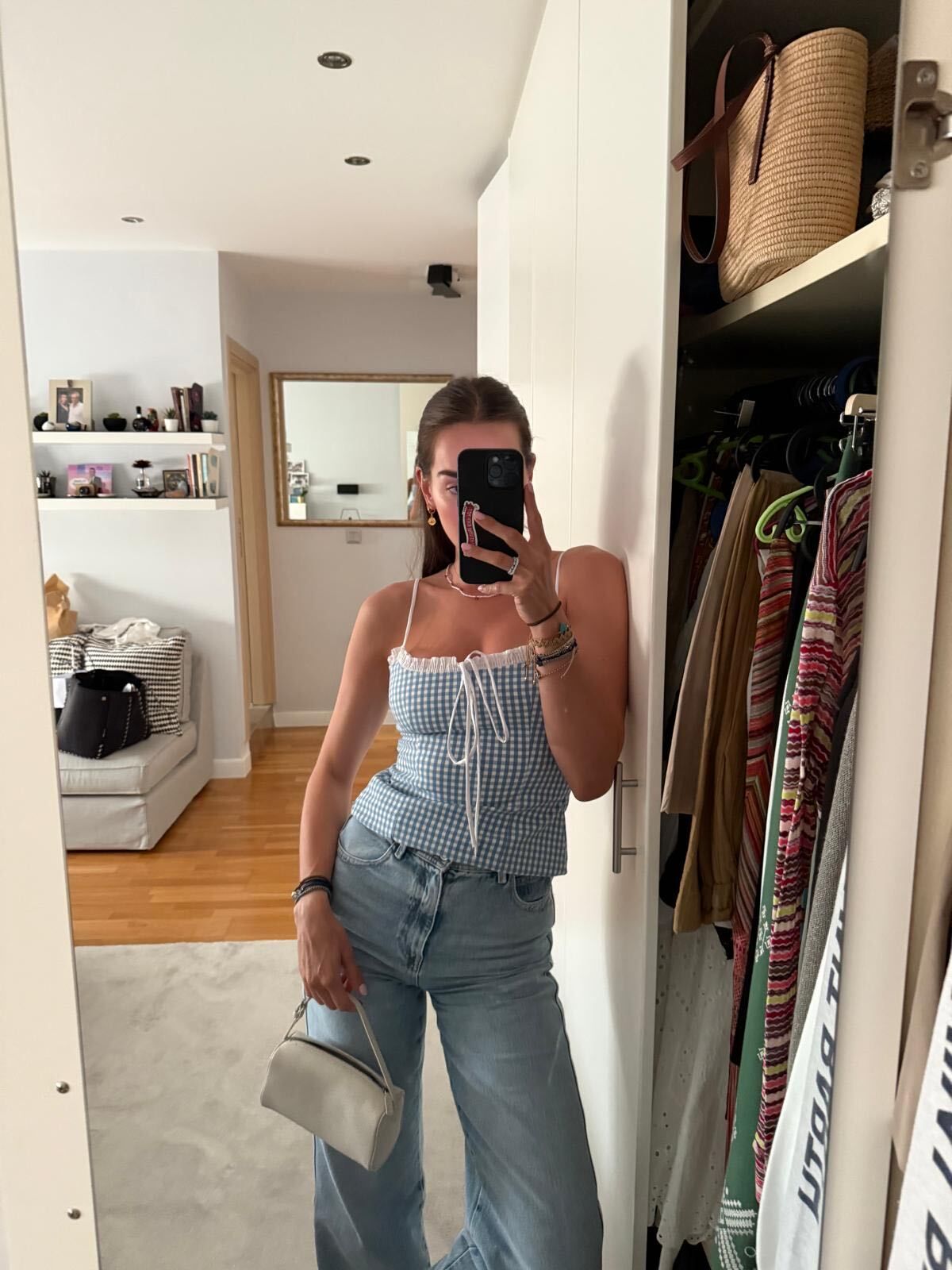 Woman with brown hair wearing gingham top and blue jeans and white bag taking a mirror selfie.