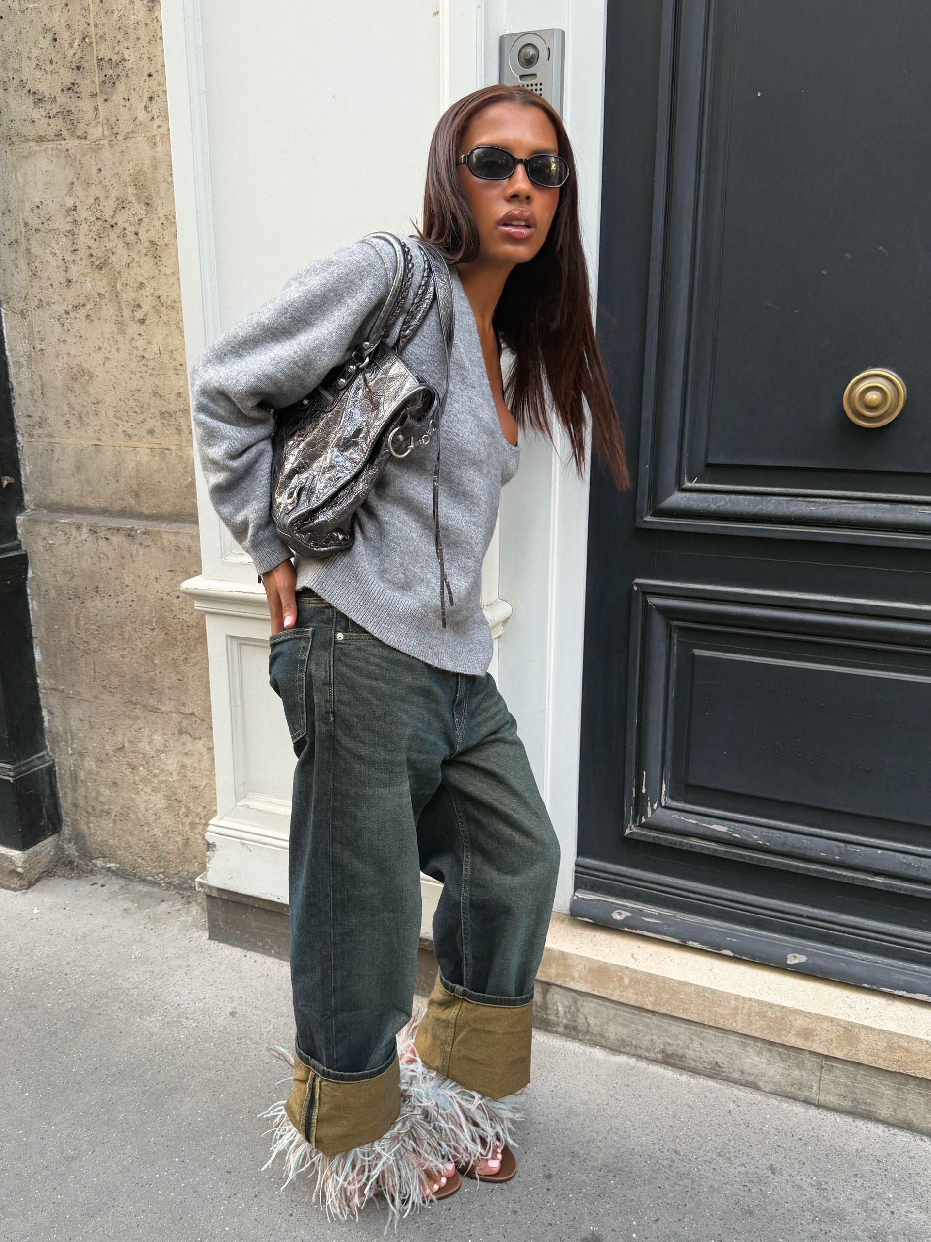 An image of @mv.tiangue wearing cuffed jeans with a grey knit and feathered shoes.