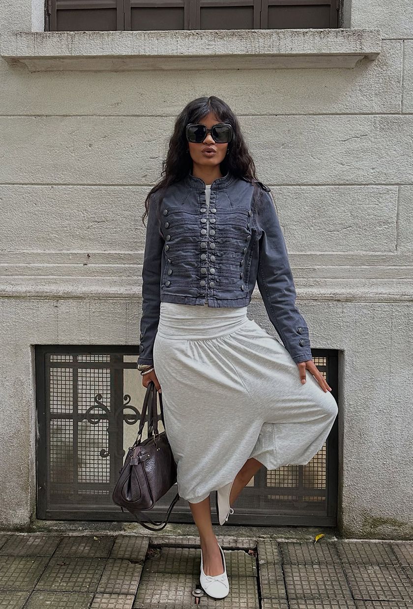A woman wearing a dark gray Napoleon jacket with a gray tank, gray balloon pants, ballet flats, a handbag, and oversize sunglasses.
