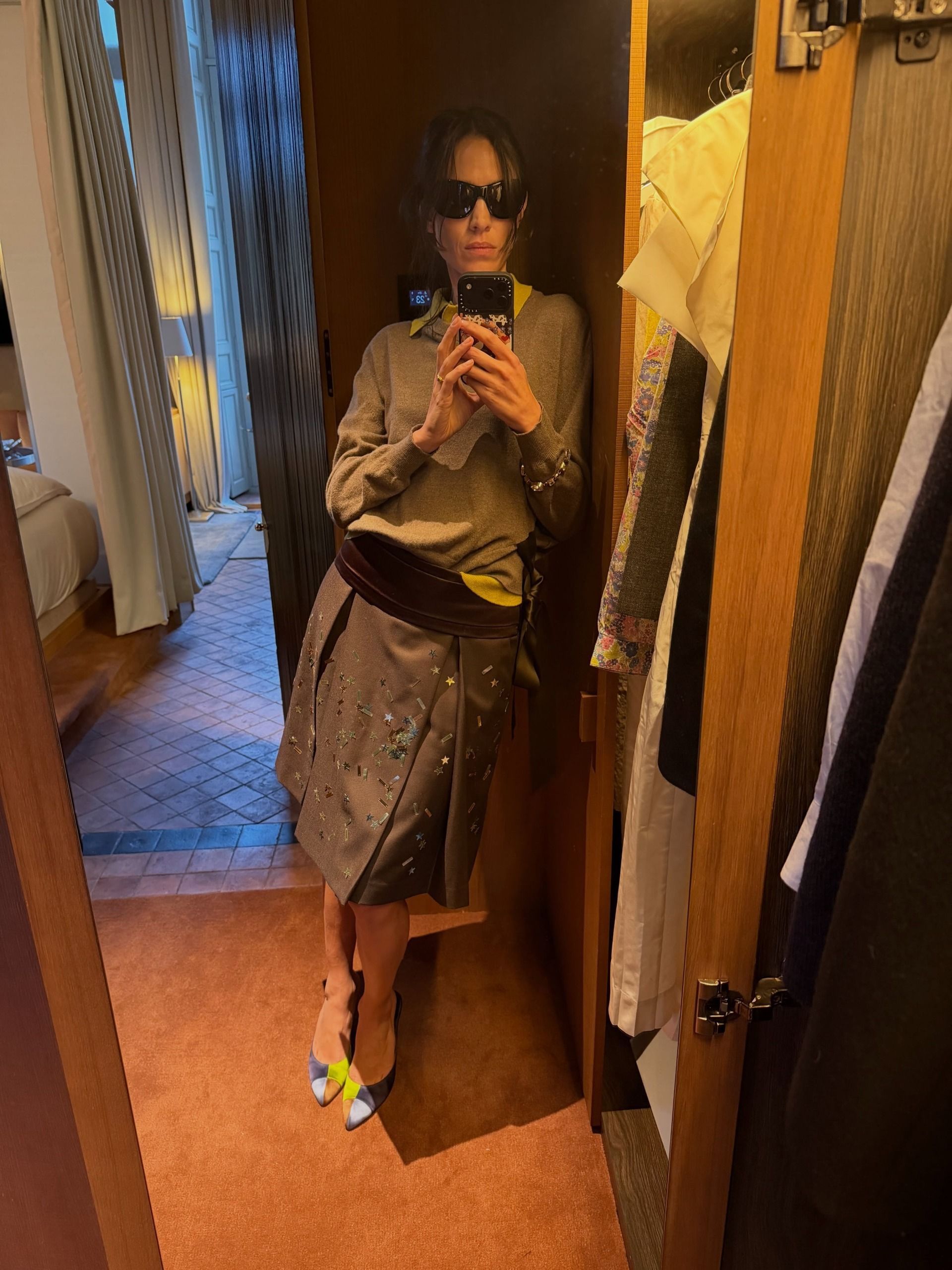 Alexa Chung takes a mirror selfie wearing a knee-length pleated skirt with a grey jumper, a yellow shirt and a purple cummberbund.
