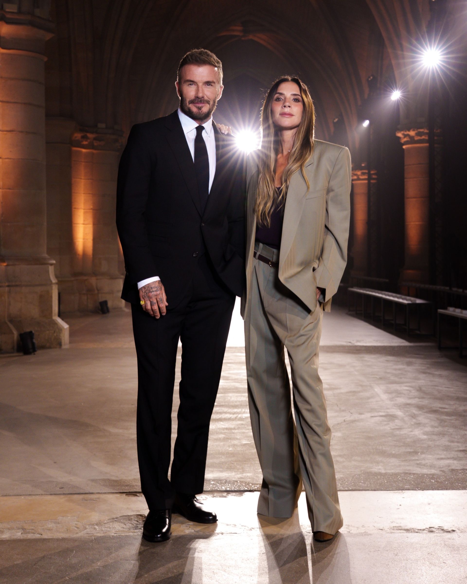 Victoria Beckham poses at the Victoria Beckham SS27 runway show with David Beckham. She wears a grey suit with brown pointed-toe heels.