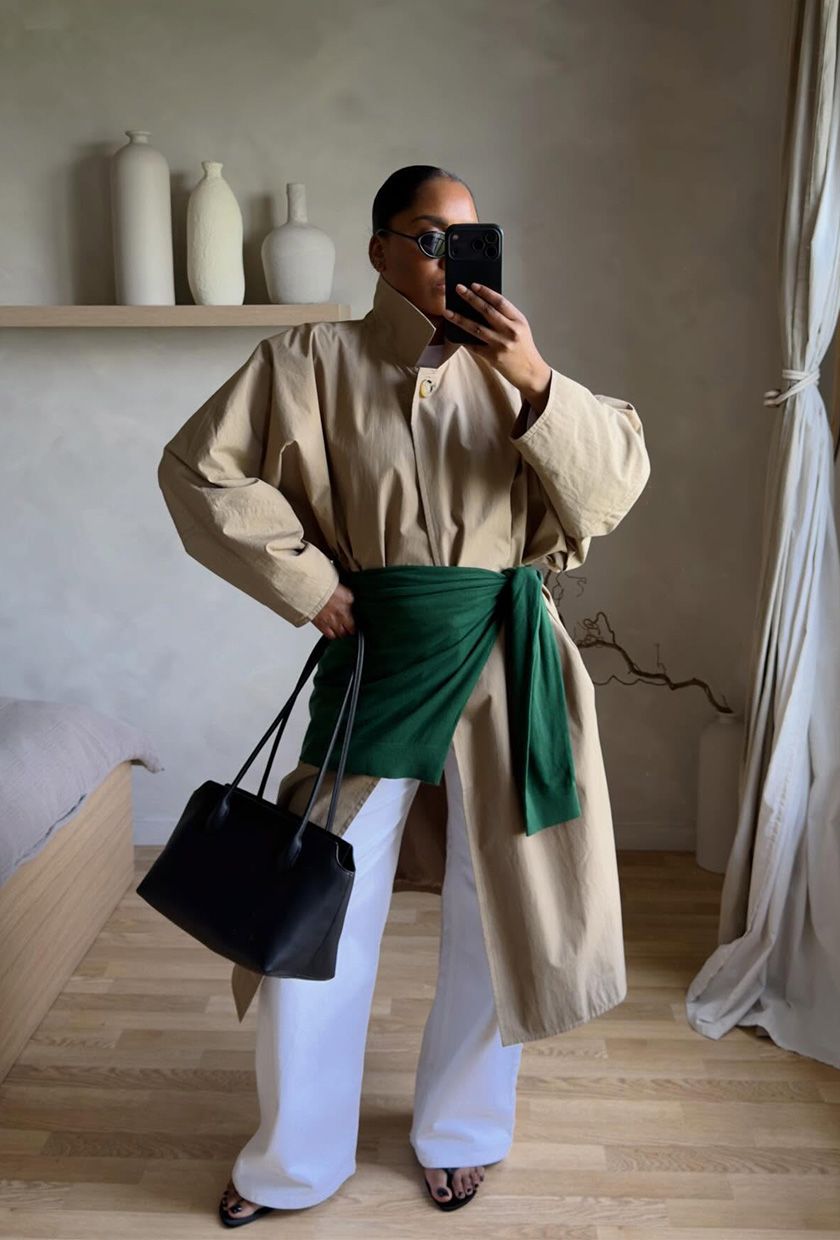 A woman wearing a tan trench coat with a green sweater tied around the waist, styled with white jeans, black flip-flops, and a black bag.