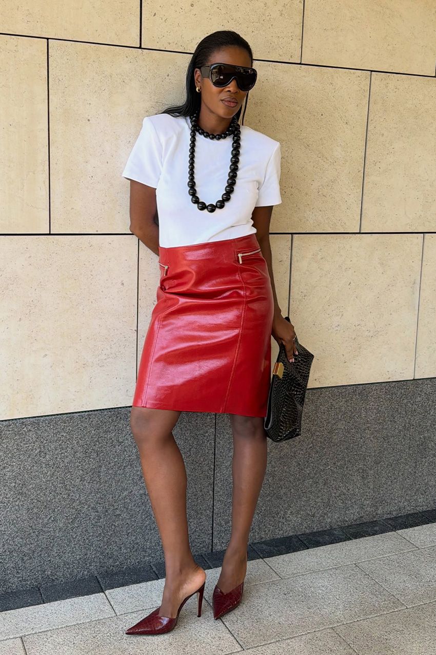 A woman wearing a white T-shirt tucked into a red leather pencil skirt, styled with a long black necklace, red heeled mules, a black clutch, and sunglasses.