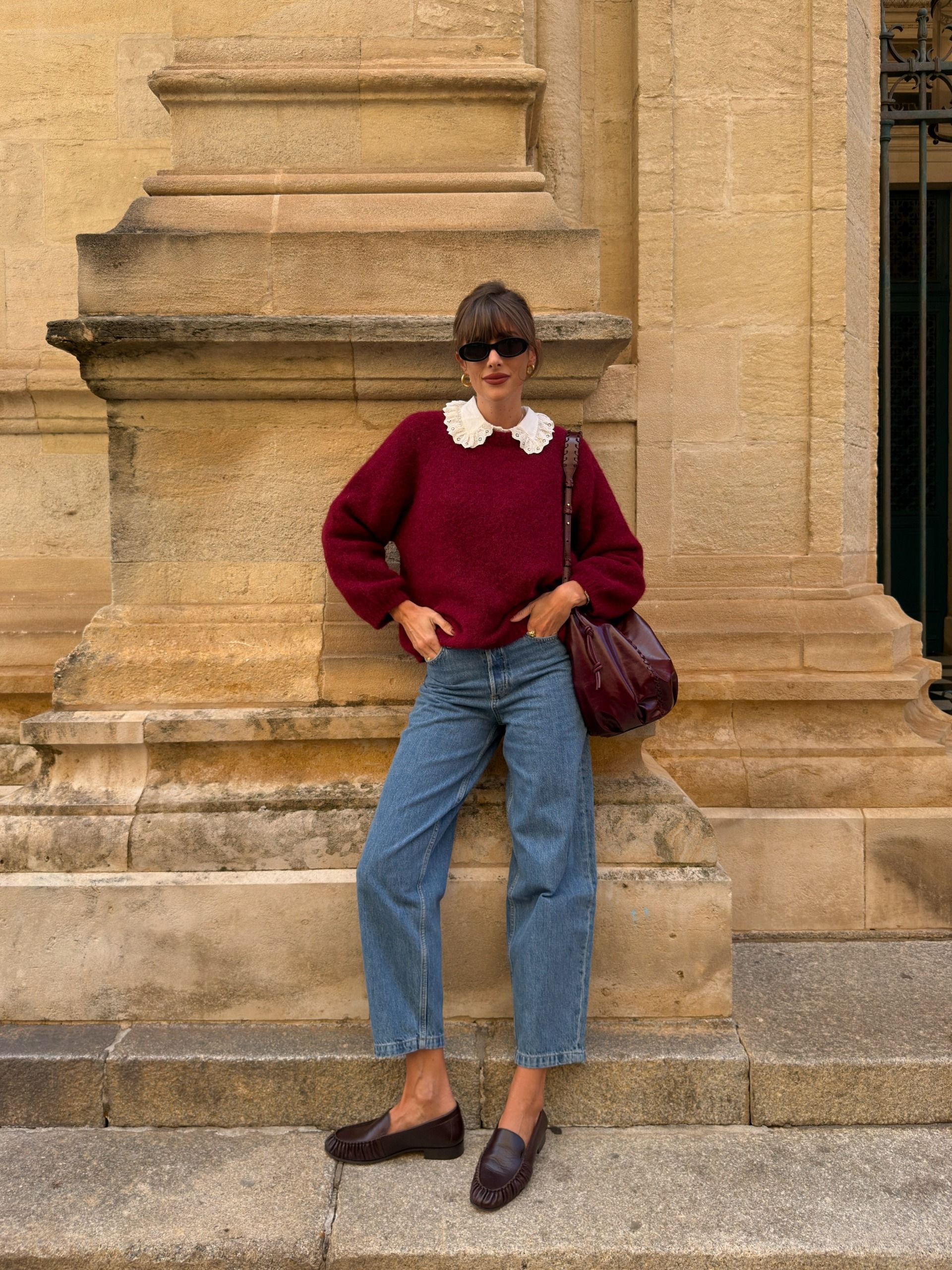 Influencer @juliesfi poses for a photo wearing a burgundy knit with blue jeans, brown loafers and a burgundy bag.